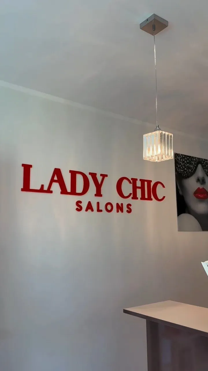 Sarkans uzraksts „Lady Chic Salons” uz gaišas sienas un kristāla lampa uzgaidāmajā zonā